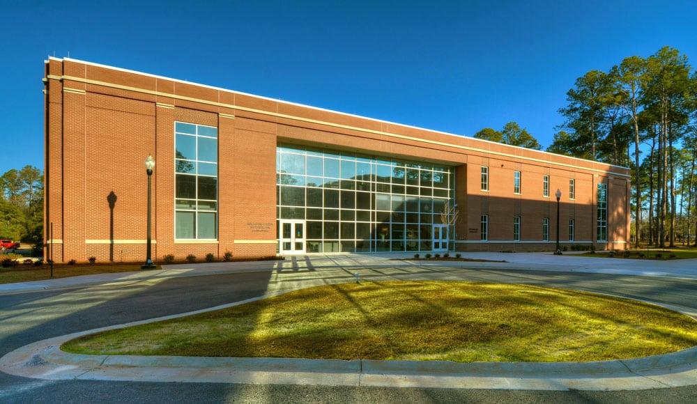 A two-story brick building with expansive glass windows, set among a driveway, neatly trimmed shrubs, and tall trees beneath a clear blue sky.