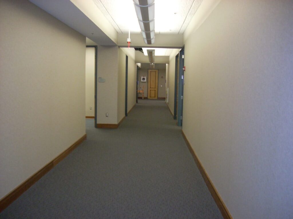 A quiet, empty hallway with beige walls, gray carpet, and a wood-trimmed baseboard. Fluorescent lights run along the ceiling, and there are doors and signage visible at the end of the hallway.