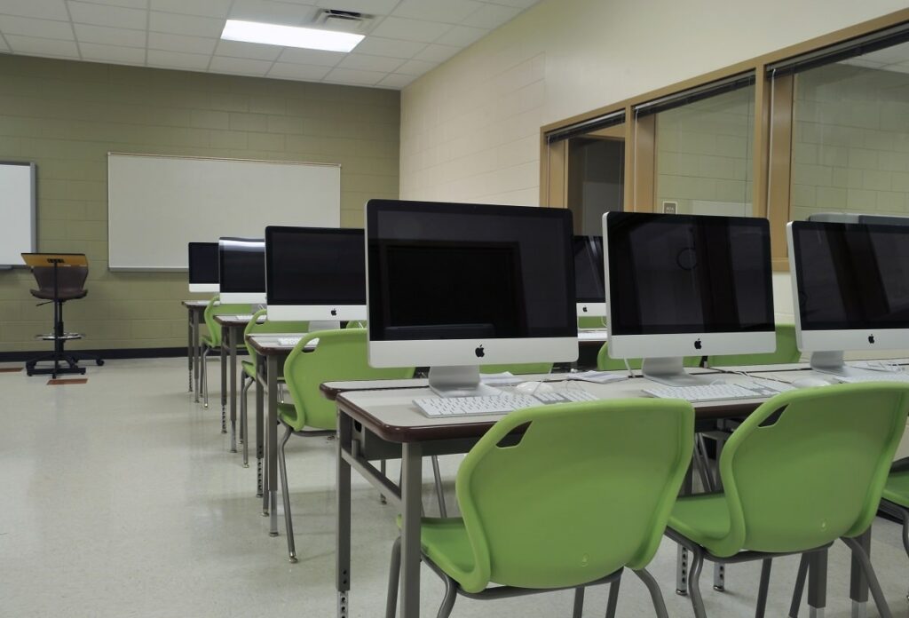A classroom contains rows of Apple desktop computers on desks, each with a green chair. At the front, there is a whiteboard on beige walls and a lectern.