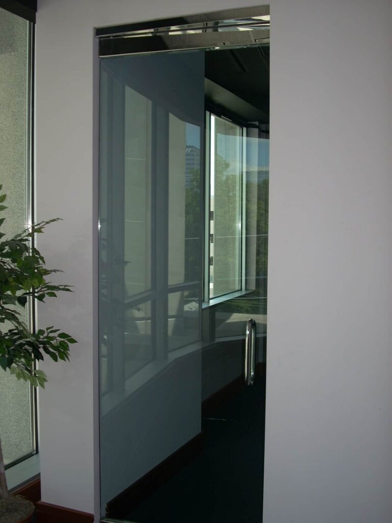 A nearly invisible glass door is open in an office space, blending with the surrounding glass windows and white walls. To the left, there’s a leafy plant.