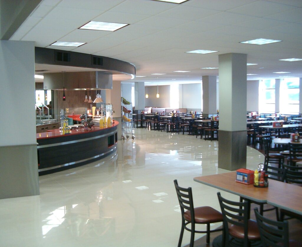A spacious cafeteria with many empty tables and chairs, bright lighting, and a serving counter on the left. Large windows let in natural light, creating a clean, modern atmosphere.