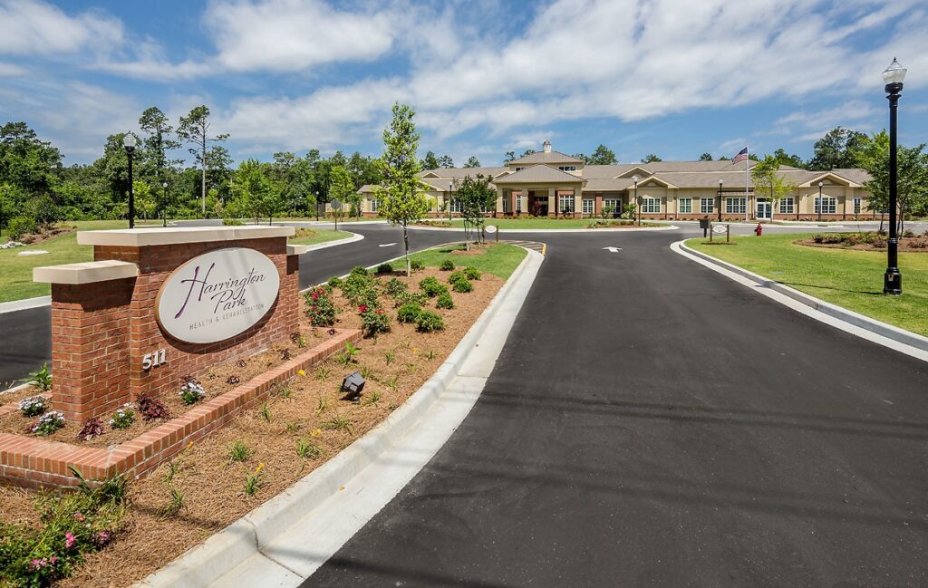 A paved driveway leads to a large, single-story building surrounded by grass and trees. A brick sign at the entrance reads Harrington Park. The sky is partly cloudy.