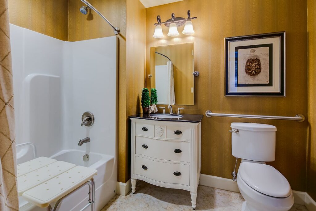 A bathroom with a white bathtub and shower, a safety seat, a toilet with grab bars, a white vanity with a mirror, and gold walls with a framed picture. Three lights are above the mirror, and a small plant is on the vanity.