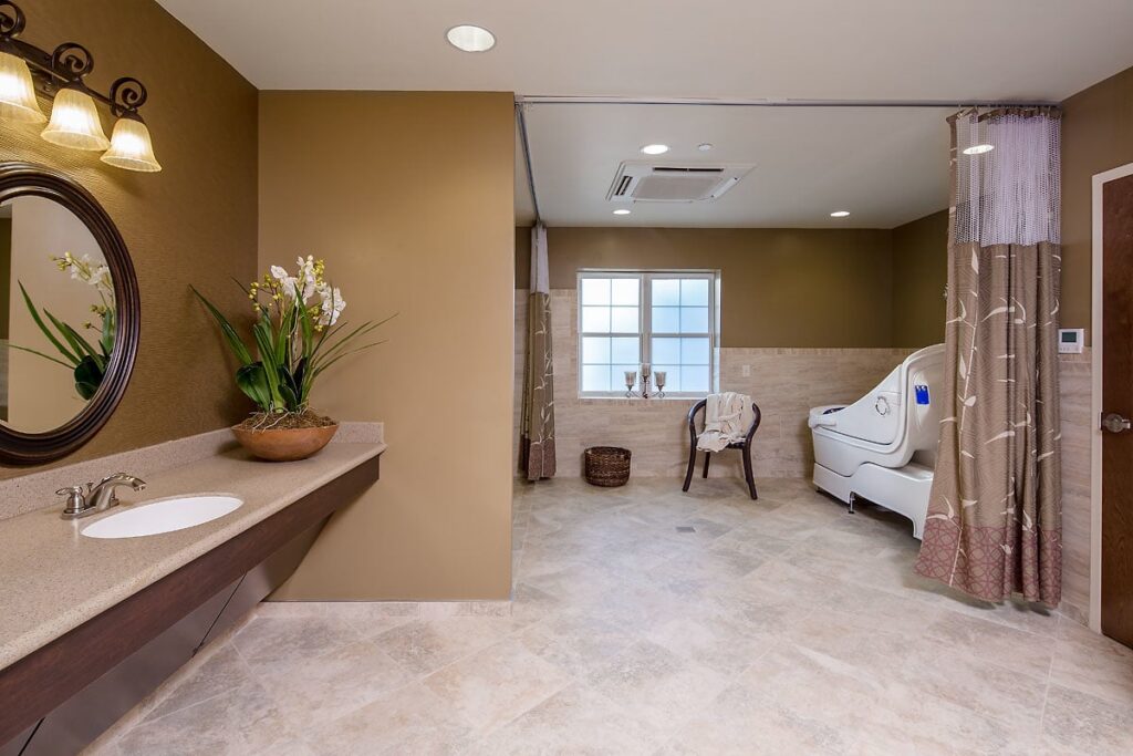 A spacious bathroom featuring beige walls and floor, a round mirror above the sink, a potted plant, and a walk-in bathtub situated near a window with a curtain for separation.