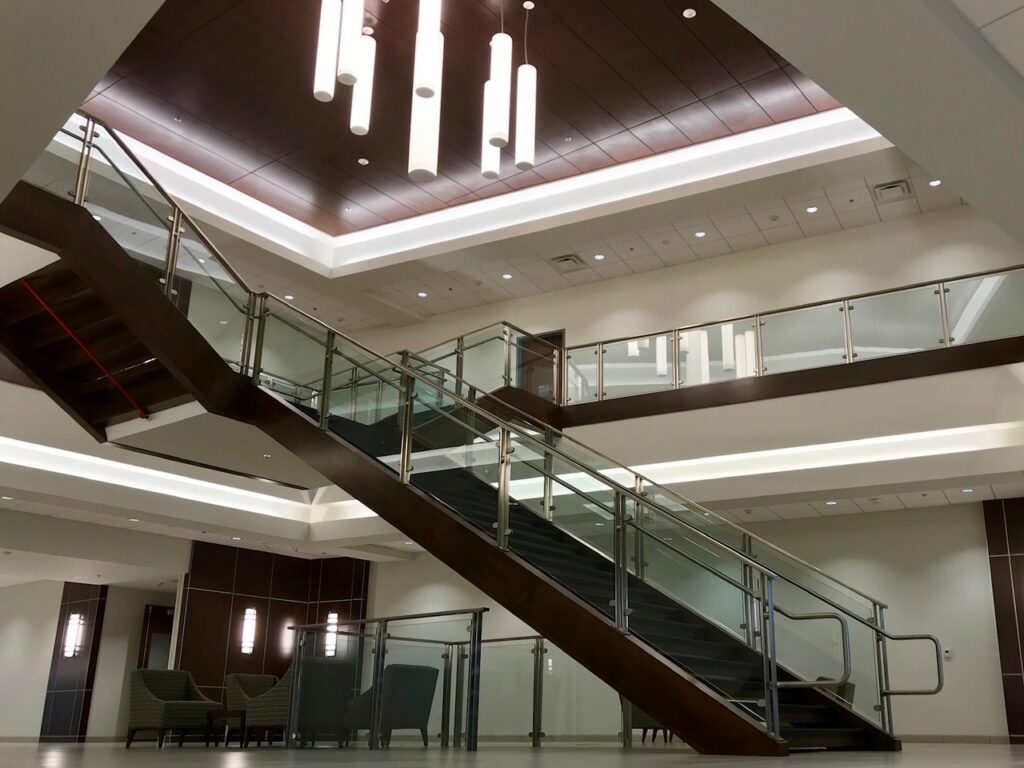 Modern interior featuring a prominent staircase with glass railings ascending to an upper level. Contemporary light fixtures are suspended from the ceiling, and multiple chairs are arranged in a seating area beneath the stairs.