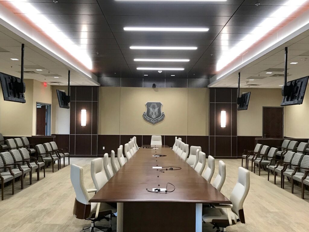 A spacious conference room featuring a long wooden table surrounded by white chairs, multiple wall-mounted monitors, and rows of extra seating. A prominent emblem is visible on the far wall beneath bright ceiling lighting.