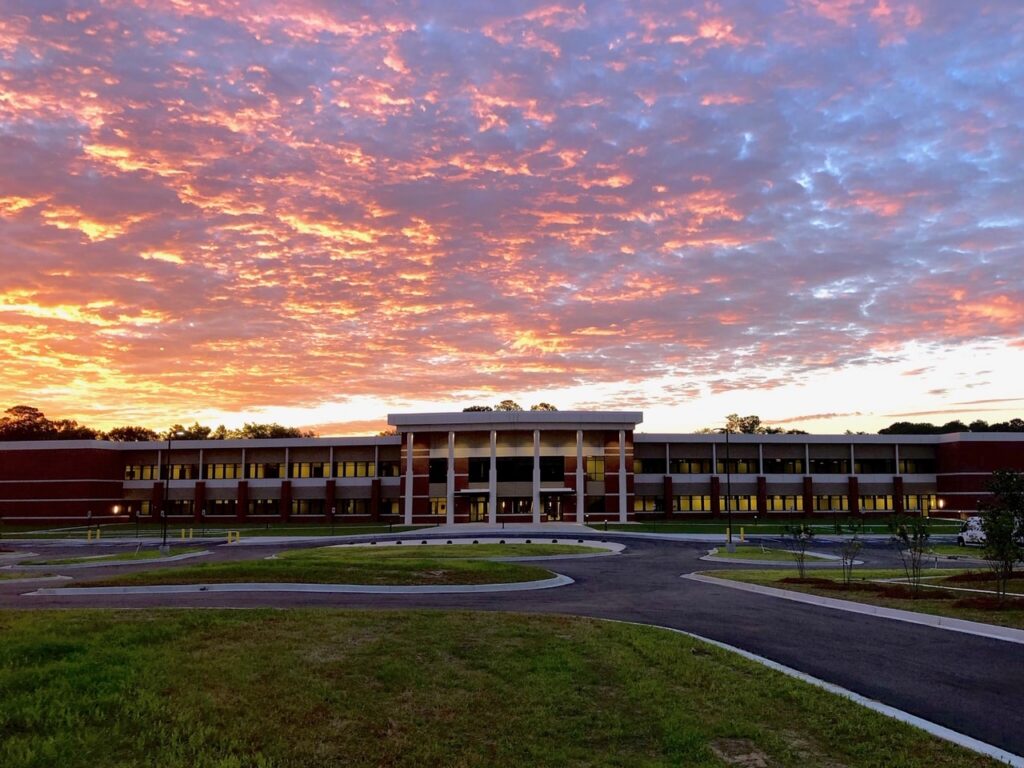 A modern two-story brick building with columns stands under a vibrant orange and purple sunset sky. In the foreground, there’s a curving driveway and green lawn.