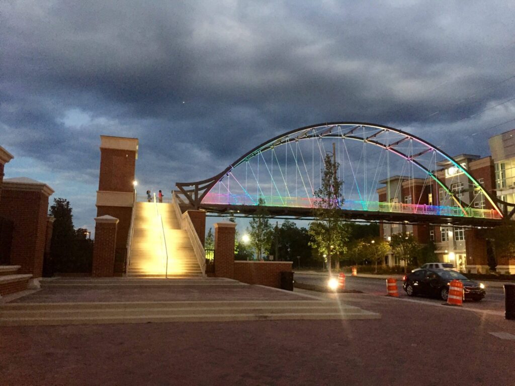 A pedestrian bridge illuminated with rainbow-colored lights stretches over a street at dusk, featuring glowing stairs ascending on the left and buildings visible in the background.