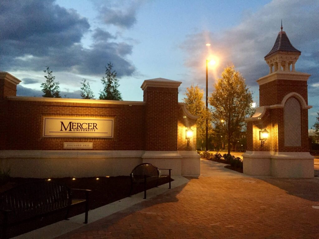 Brick entrance with Mercer University sign, illuminated by lamps, flanked by two towers. Trees and benches line the walkway; sky is dusky with clouds and a bright streetlight in the background.