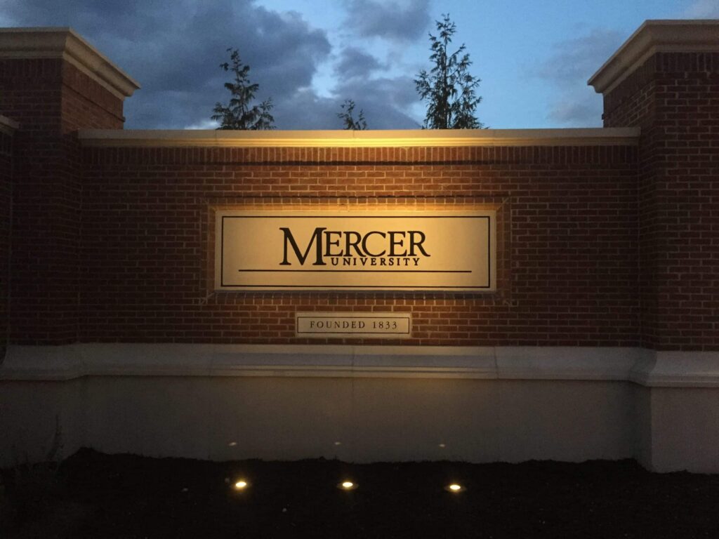 A brick wall sign for Mercer University is illuminated at dusk. The words Founded 1833 appear beneath the main sign. Trees and a cloudy sky are visible in the background.