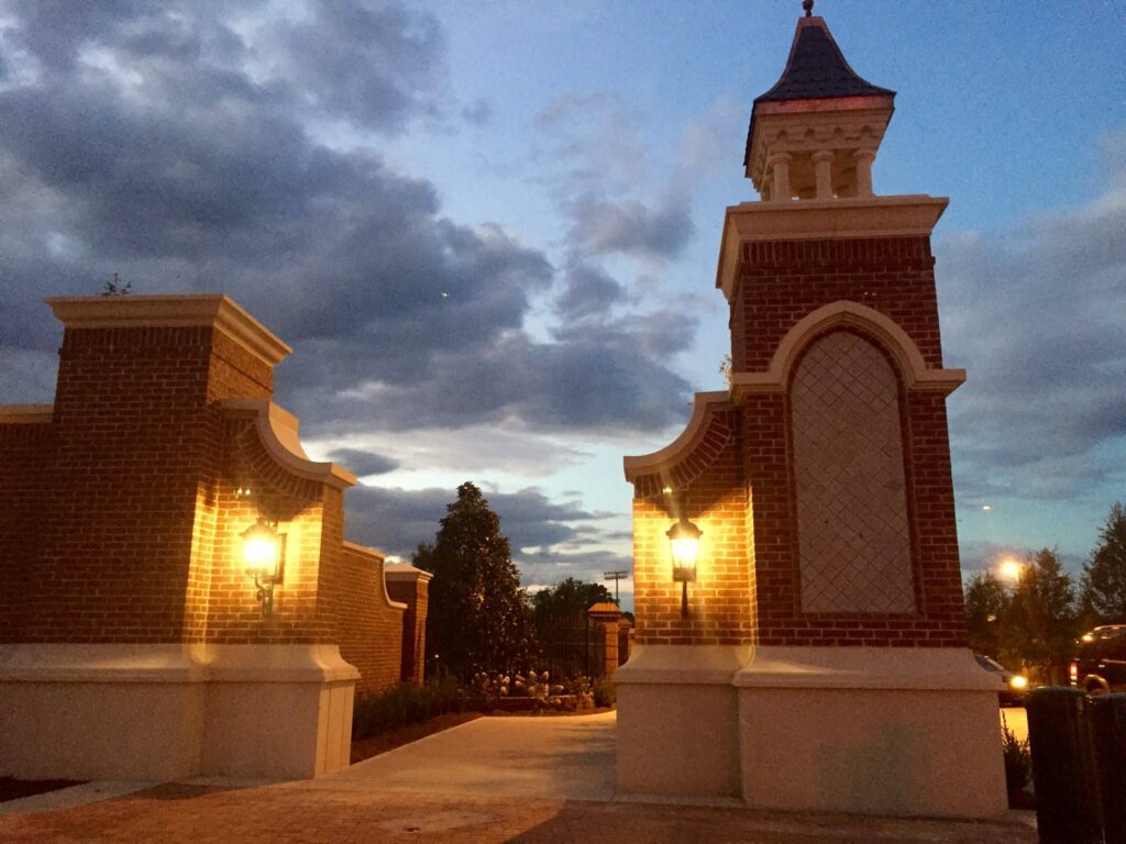 Brick entrance gates with decorative lamps glow at dusk, set against a cloudy sky and silhouetted trees in the background. The atmosphere is serene and welcoming, with warm illumination emphasizing the architectural features.