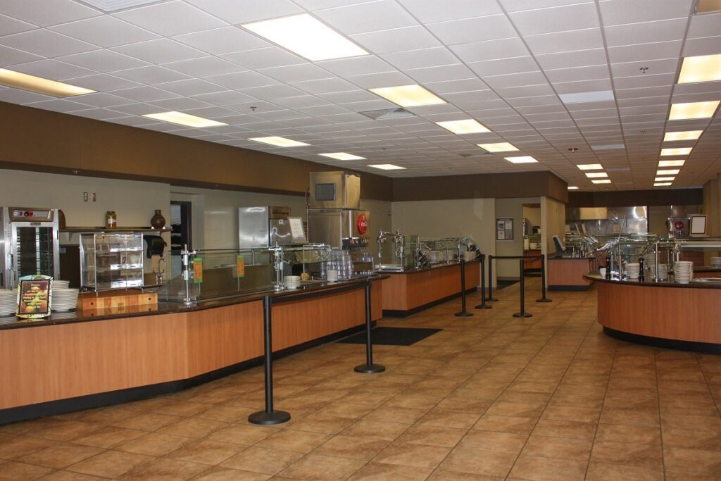 An empty cafeteria featuring buffet-style serving counters, illuminated by overhead lighting with a tiled floor. There are no people present. Behind glass partitions on the counters, food displays and utensils can be seen.