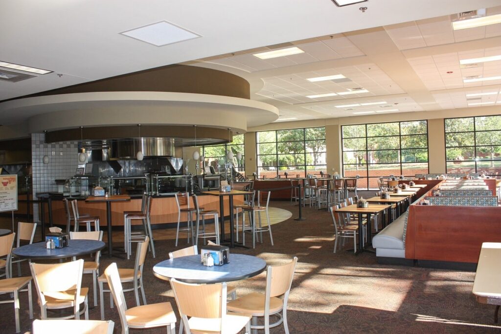 Bright, modern cafeteria featuring empty tables and chairs, large windows streaming sunlight inside, and a food serving area with metal counters and stools. Trees can be seen outside the windows.