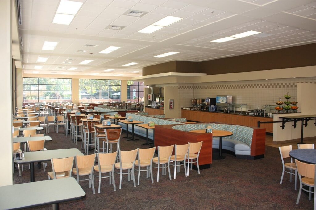 A spacious, well-lit cafeteria featuring rows of light wooden tables and chairs, cushioned booth seating, a self-serve drink station, and large windows filling the space with natural light.