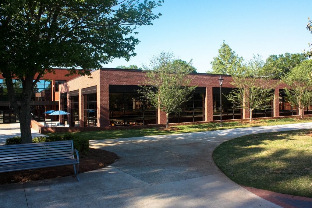 A brick building with large windows stands behind some trees and a curved sidewalk. In the shaded courtyard, a metal bench and a blue umbrella shading outdoor tables can be seen. The sky above is clear and blue.