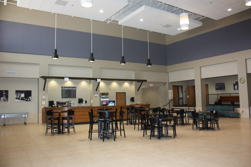 Spacious cafe featuring several black tables and chairs, a counter topped with menu boards, hanging lights, and neutral-colored walls. The interior is decorated with a few framed photos and a clock.