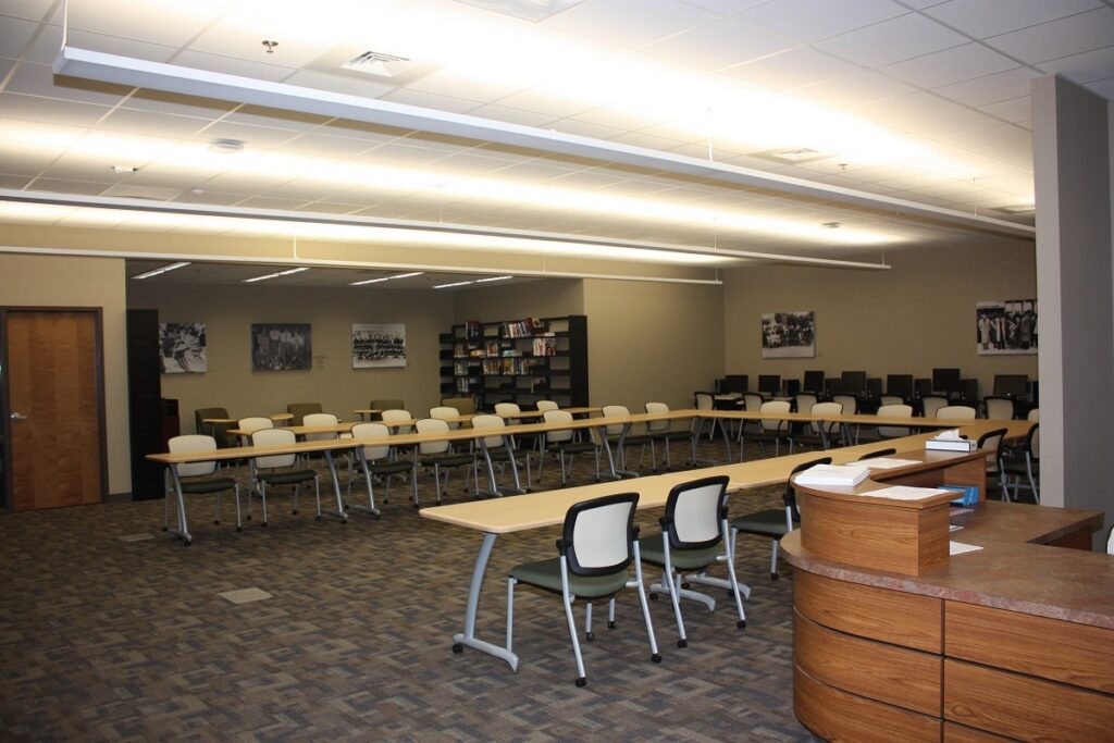 A spacious, well-lit library or classroom featuring rows of desks and chairs, bookshelves lining the wall, computers positioned in the back, and black-and-white photos displayed on the walls.
