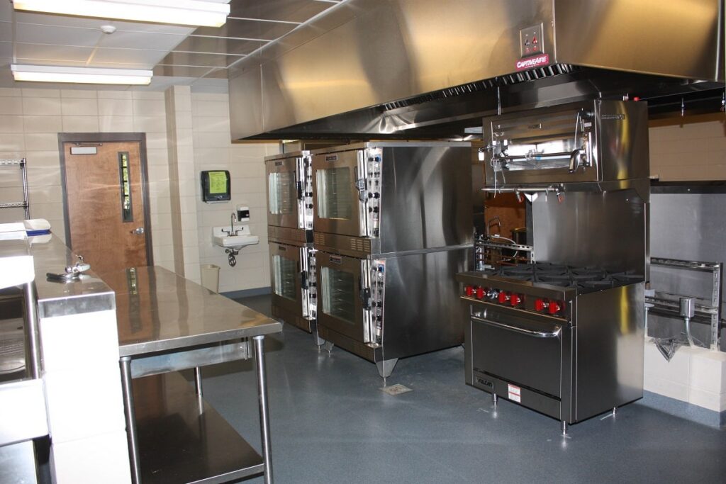 A commercial kitchen featuring stainless steel appliances such as ovens, a stove, and prep tables. The area appears clean, with a sink and a door in the background, all illuminated by bright overhead lighting.