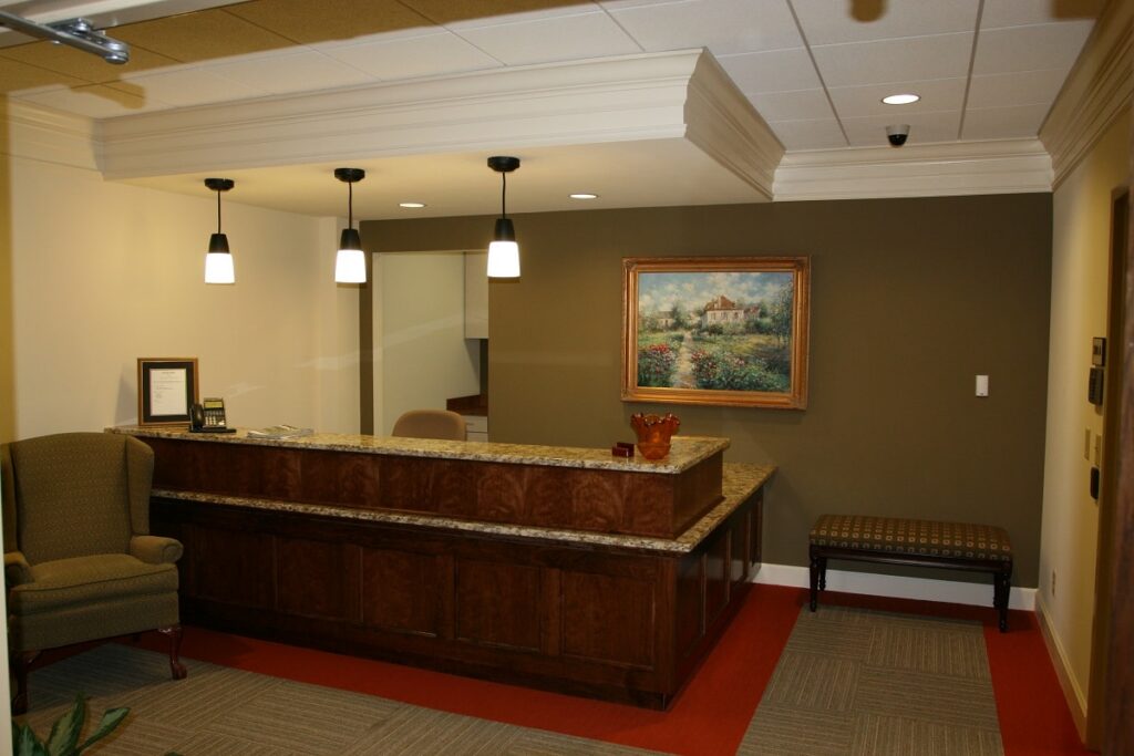 Reception area featuring a marble-top desk, three pendant lights, a framed painting on the wall, a green armchair, a cushioned bench, and neutral-toned carpeted flooring.