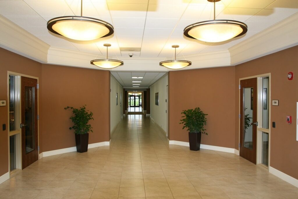 A tidy hallway features tan walls, light tile flooring, four ceiling lights, two potted plants, and multiple doors lining both sides, all leading toward a bright space at the end.