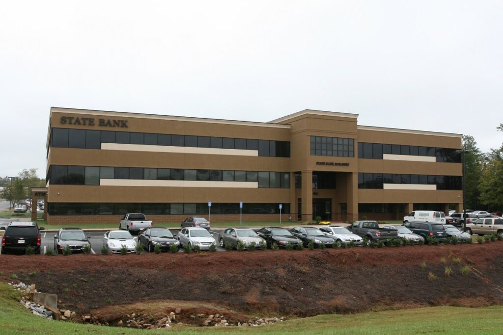 A three-story tan and brown building labeled State Bank, with many cars parked in front on a cloudy day. Grass and a small ditch are visible in the foreground.