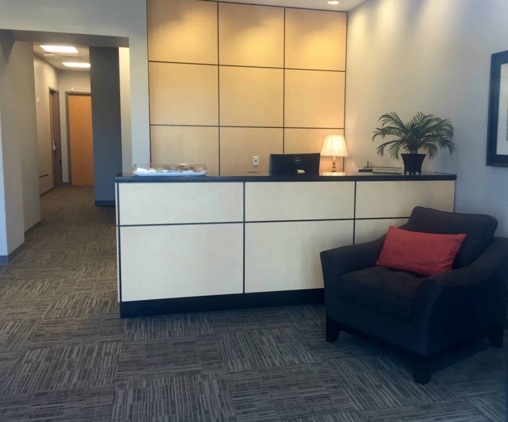Reception area featuring a light wood desk, a small lamp, a potted plant, and a dark armchair with a red pillow. A hallway is visible to the left, wall art hangs on the right, and neutral grey carpeting covers the floor.