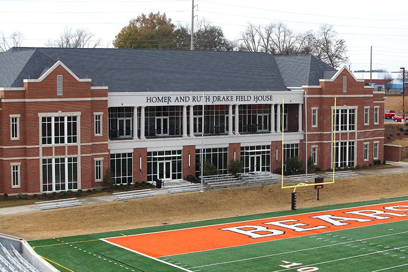 A large brick building labeled Homer and Ruth Drake Field House overlooks a football field with orange end zones and BEARS written in the end zone.