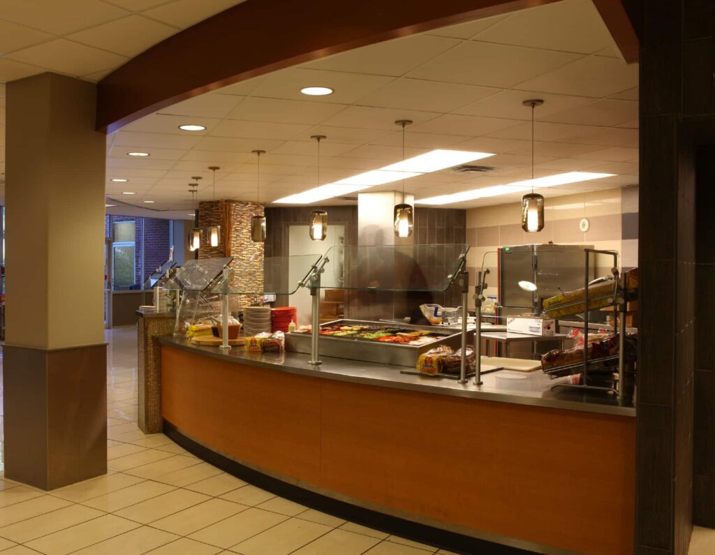 A cafeteria counter with overhead lights showcases trays of food and baked goods behind a glass barrier. Stacks of plates and loaves of bread are present, set against a background of tiled flooring and neutral-colored walls.
