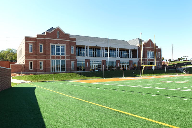 A spacious brick school building with tall windows rises behind a green football field, its yard lines clearly marked, all beneath a bright blue sky.