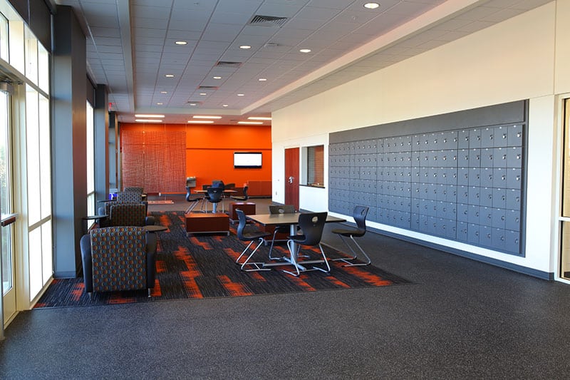 Modern lounge area featuring chairs, tables, and lockers along the wall. Large windows allow natural light to fill the space. The walls are painted orange and gray, and the floor is covered with dark carpet and patterned rugs.