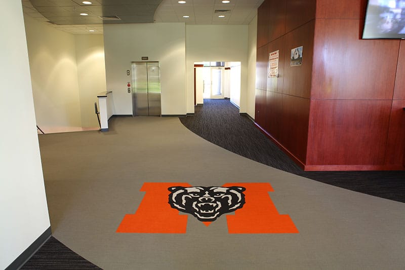 A hallway featuring an elevator, wood-paneled walls, and a large orange M with a bear mascot logo displayed on the carpet.