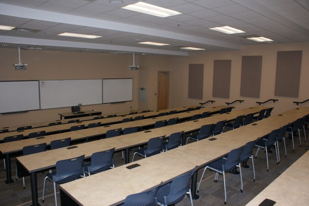 An empty classroom features rows of desks and chairs aligned toward whiteboards, a projector positioned on a table, and acoustic panels mounted on the walls. The room is brightly illuminated by ceiling lights.
