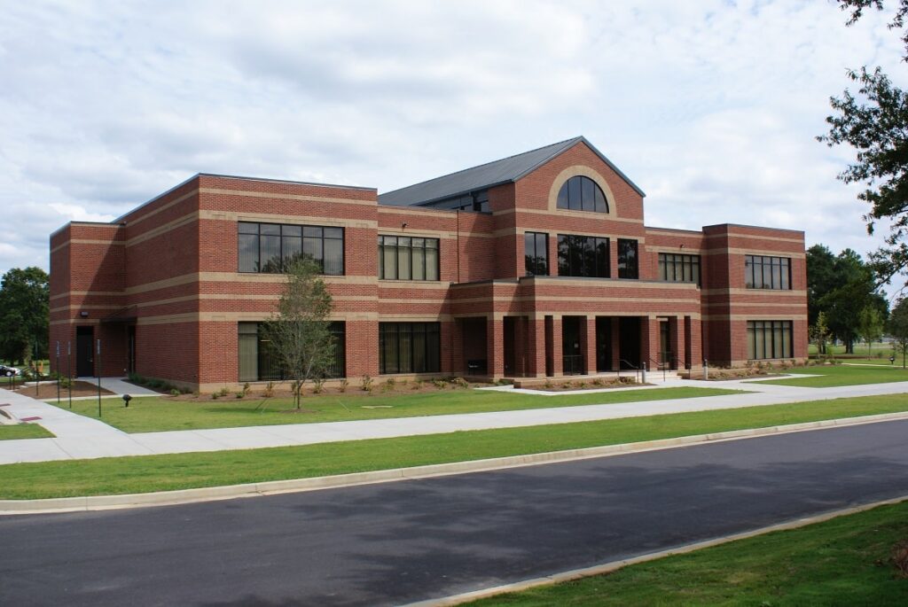 A sleek, contemporary two-story red brick building features expansive windows and is bordered by well-kept green lawns and sidewalks, set beneath a partly cloudy sky.
