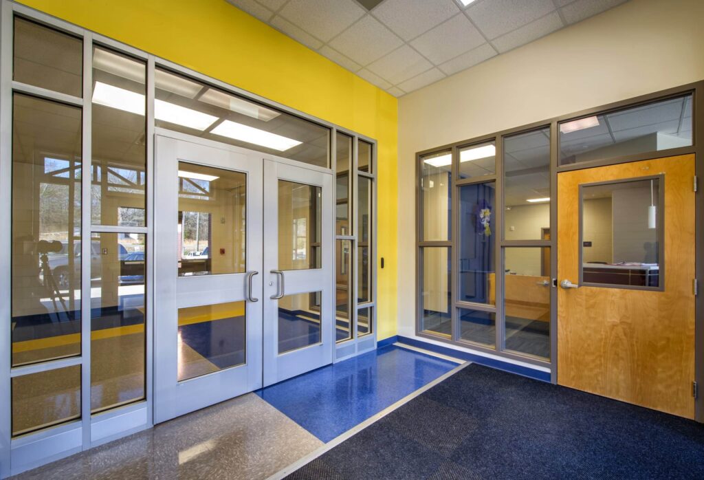 School hallway featuring yellow and beige walls, blue and gray flooring, glass doors, and a wooden door with windows. Sunlight streams in from outside, making the space look bright and inviting.