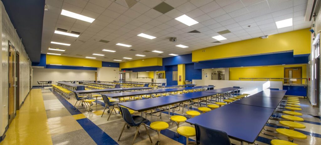 A brightly lit school cafeteria features blue tables, yellow round stools, and geometric blue and yellow wall accents; the space is empty and orderly.
