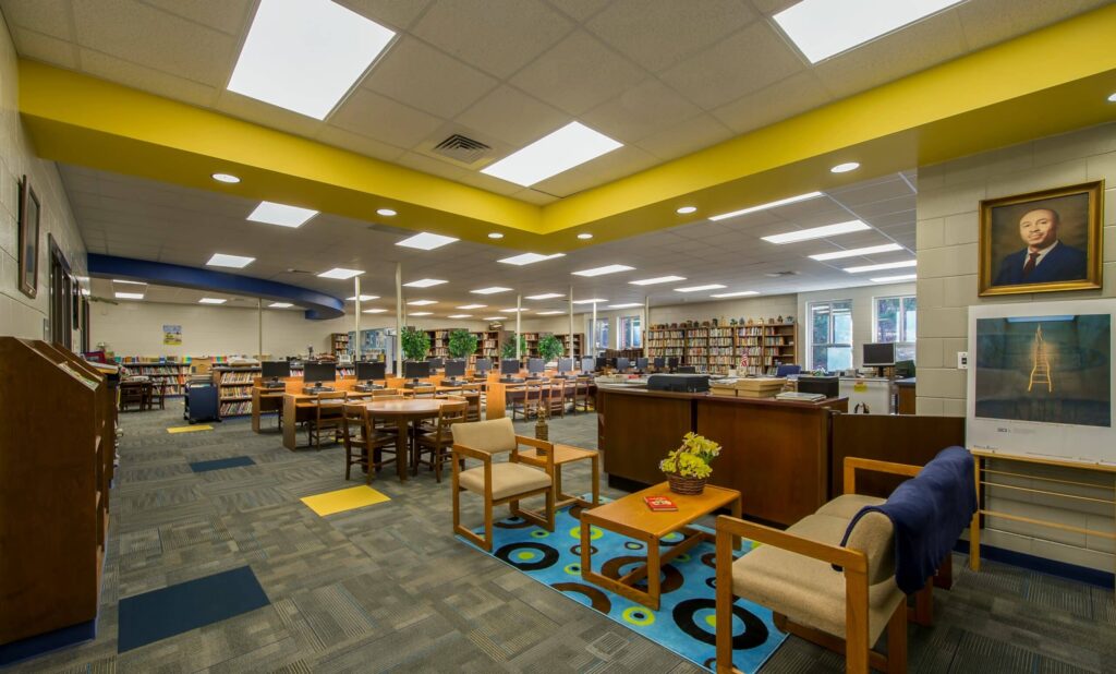 A spacious library with bookshelves, tables, and chairs. There’s a seating area with two armchairs and small tables, plus a portrait and poster on the wall. Bright lights and windows create a well-lit, inviting atmosphere.