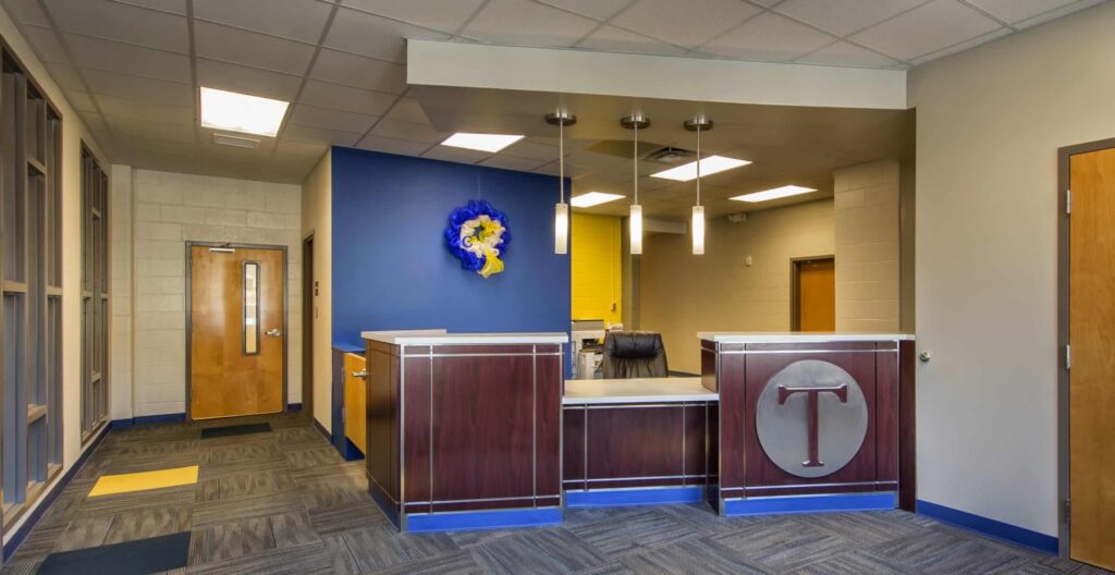 Modern school office with blue and yellow walls, wood reception desk with a large T emblem, overhead lights, and multiple doors. A chair is behind the desk and a decorative wall piece hangs on the blue wall.