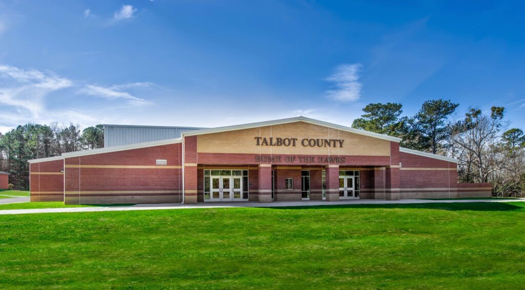 A brick school building with Talbot County Home of the Tigers above the entrance, surrounded by green grass and trees under a blue sky with light clouds.
