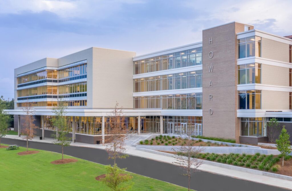 Modern four-story school building with large windows and Howard written vertically on the front wall. The entrance has stairs and landscaping, and the area in front features a lawn and paved walkway.