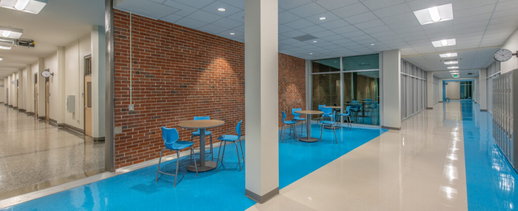 A modern school hallway featuring blue and white floors, blue chairs, and small round tables. Lockers are lined up on the right side, while brick and glass walls with bright lighting add a contemporary touch.