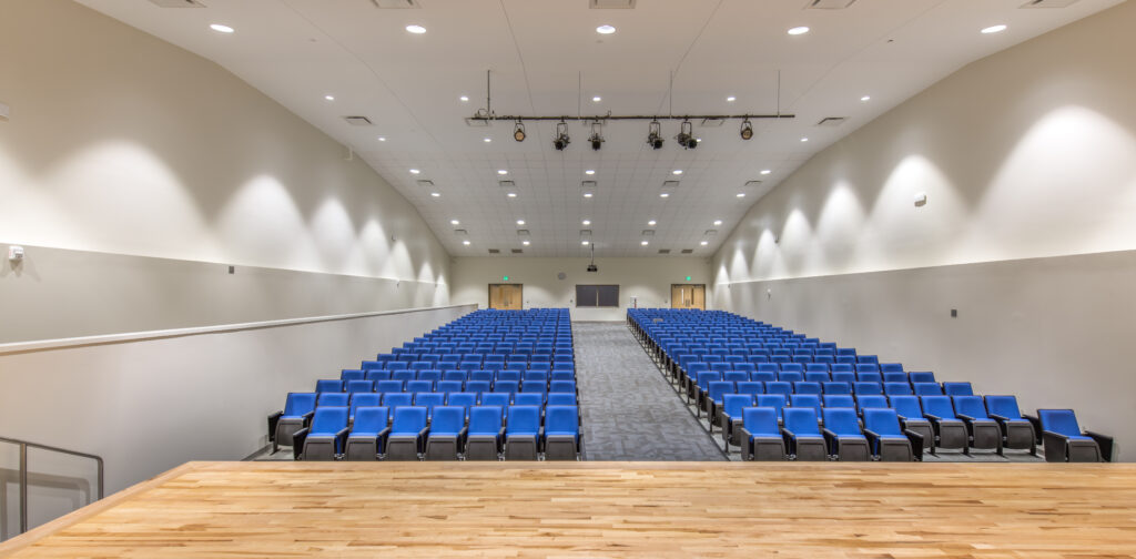 A view from a wooden stage looking out at an empty auditorium featuring rows of blue seats, light-colored walls, and a high ceiling with lights and speakers.