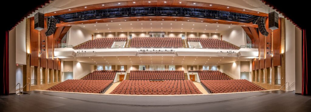 View from a theater stage looking out at the audience area, featuring multiple rows of empty brown seats, balconies, and contemporary architectural elements illuminated by warm lighting.
