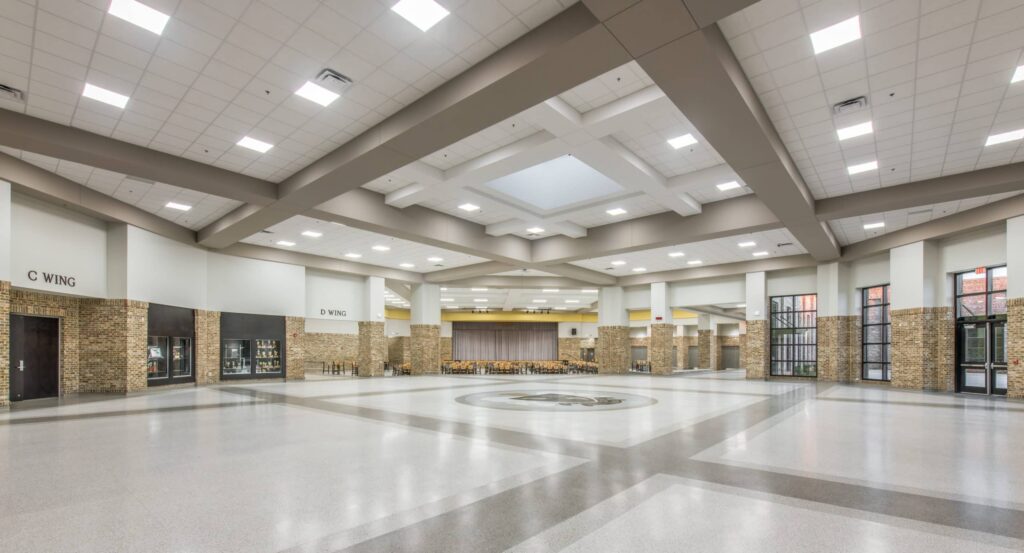 A spacious, modern school lobby with high ceilings, large windows, brick accents, and polished floors. Signs indicate C Wing and D Wing. Rows of chairs are visible at the back of the area.