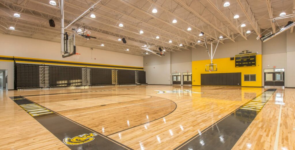 Empty indoor basketball gym featuring polished wood floors, a scoreboard, basketball hoops, bleachers, and yellow and black accents on the walls. Bright overhead lights illuminate the spacious court.