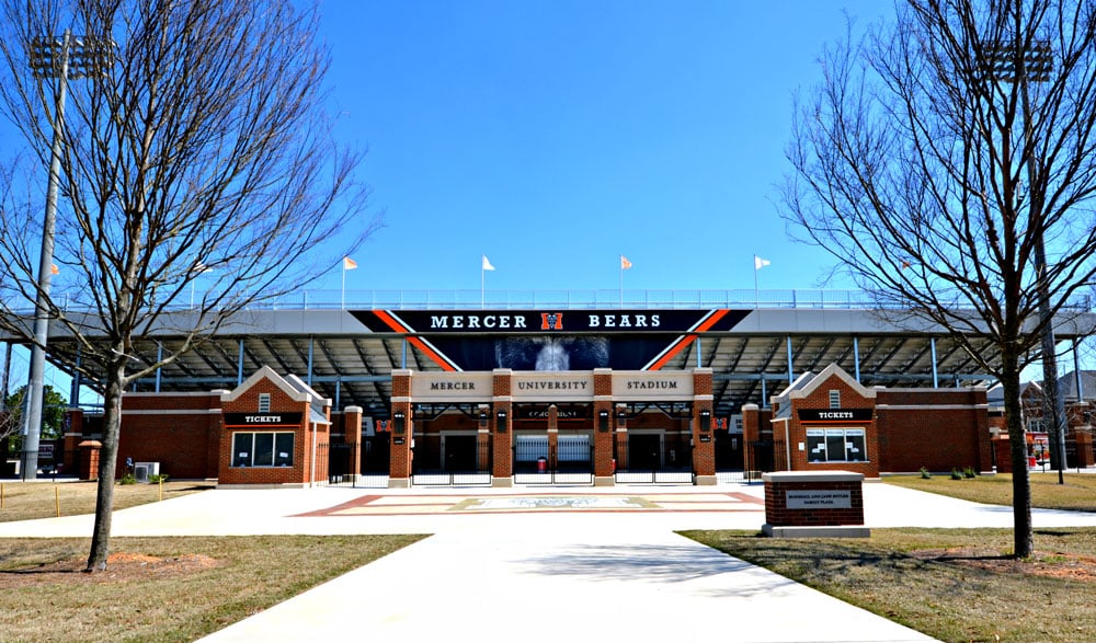 Entrance to Mercer University Stadium with brick ticket booths, leafless trees, and Mercer Bears sign above the stands on a sunny day. Sidewalk leads up to the entrance.