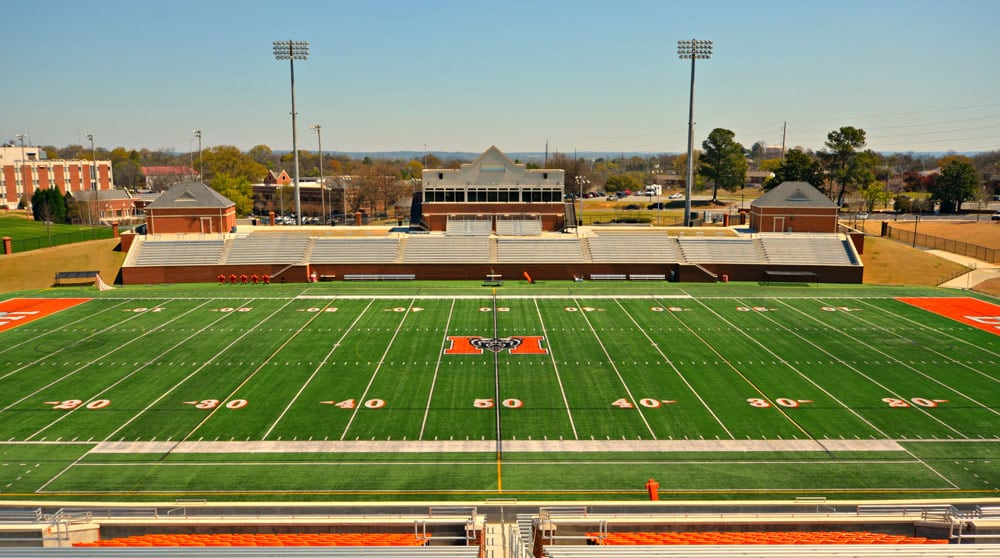 Empty football field with green artificial turf, stadium seating on both sides, orange end zones, and lights overhead under a clear blue sky. A large M logo is at the center of the field.