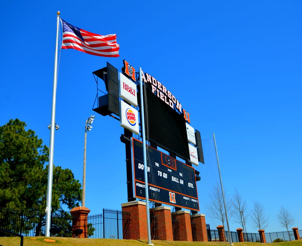 A large scoreboard at Anderson Field stands beneath a clear blue sky, with an American flag waving nearby and trees with bare branches visible in the background.