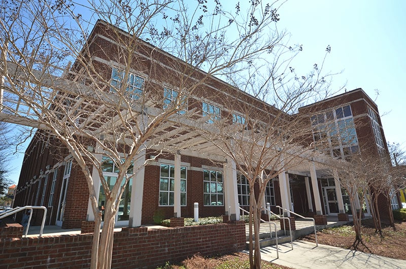 A contemporary brick building featuring expansive windows and a white pergola at its entrance, set among bare trees and neatly landscaped walkways under bright sunshine.