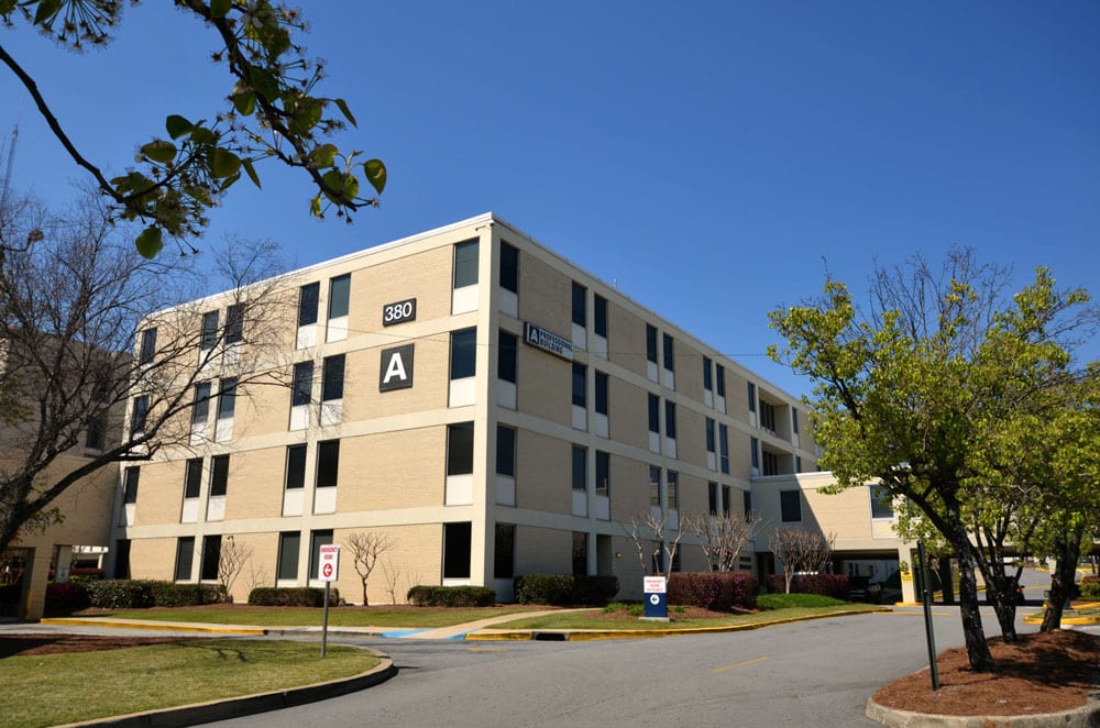 A four-story beige building labeled A and 380, with several trees and a circular driveway in front, under a clear blue sky.