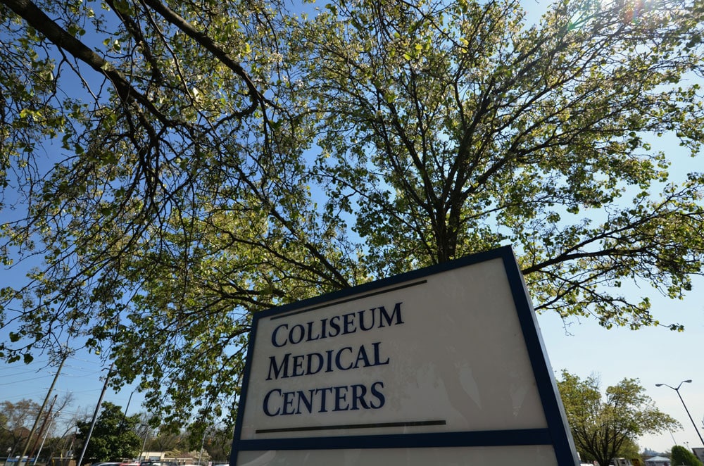A sign reading Coliseum Medical Centers stands in front of a leafy tree on a sunny day, with branches and blue sky visible in the background.